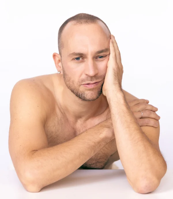 Nachdenklicher Mann mit kurzem Haar und nacktem Oberkörper, der sich mit einem Arm auf einen Tisch stützt und die andere Hand an die Wange legt