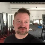 Ein Mann in einem schwarzen T-Shirt, mit Schnauzbart vollem rötlichem Haar steht in einem goßen Raum voller BDSM-Möbeln und anderen Gegenständen und schaut selbstbewusst in die Kamera.